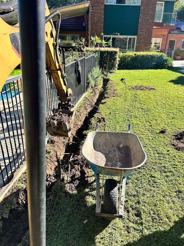Excavator cutting trench through backyard lawn near pool fence
