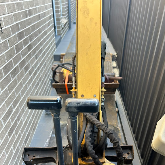 Excavator arm reaching beside a brick wall and fence line